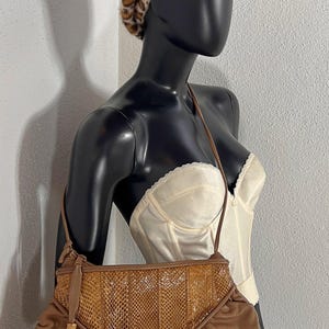 Puede incluir: Bolso de hombro de cuero marrón con un panel de piel de serpiente y una borla. El bolso se exhibe en un maniquí con un corsé color crema y una boina con estampado de leopardo.