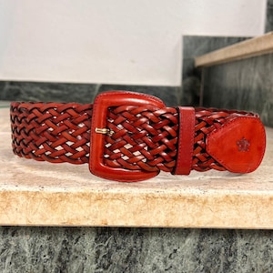 May include: A reddish-brown woven leather belt featuring a matching leather-covered buckle and a single leather keeper. The belt end is rounded and embossed with a small, detailed crest logo. The design consists of an intricate braided pattern.