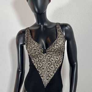 Puede incluir: Un body negro con una superposición de encaje dorado. El body tiene un escote en V y una silueta ajustada. La modelo lleva un sombrero estampado en marrón y dorado.