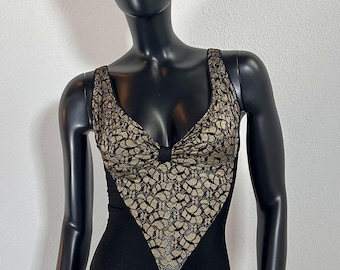 May include: A black bodysuit with a gold lace overlay. The bodysuit has a V-neckline and a fitted silhouette. The model is wearing a brown and gold patterned hat.
