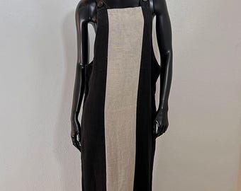 May include: A black linen dress with a central beige panel, displayed on a mannequin. The dress features buttoned straps and a long, flowing silhouette. A decorative hat with red, black, and green accents sits atop the mannequin's head.