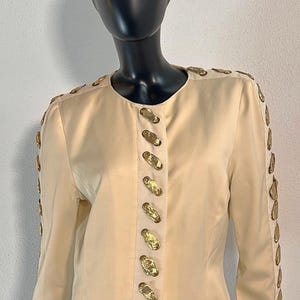 May include: A cream-colored satin jacket with gold-toned buttons and trim. The jacket has a round neckline and long sleeves.