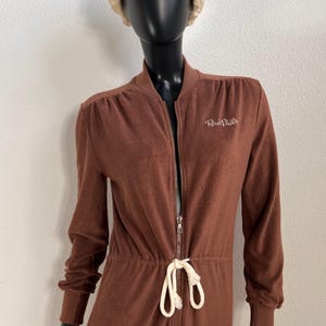 Könnte beinhalten: Ein brauner Velours-Jumpsuit mit Frontreißverschluss, einer Taille mit weißem Kordelzug und gerippten Bündchen. Im Brustbereich befindet sich der gestickte Schriftzug "René Pluill". Das Kleidungsstück wird auf einer schwarzen Schaufensterpuppe mit beiger Kopfbedeckung präsentiert.