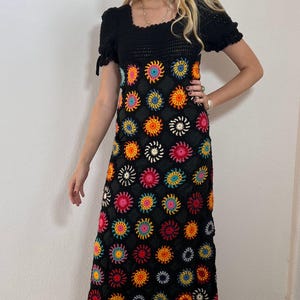 Könnte beinhalten: Ein schwarzes Häkel-Maxikleid mit bunten Blumenakzenten. Das Kleid hat einen quadratischen Ausschnitt, kurze Ärmel und ein figurbetontes Oberteil. Der Rock ist mit leuchtenden, mehrfarbigen Blumenmotiven verziert.