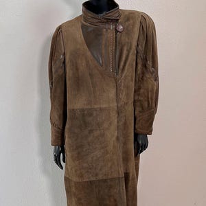 May include: A brown suede coat with a high collar and long sleeves. The coat features a patchwork design and a large button closure. A black fur hat is on the mannequin's head. The coat is knee-length.