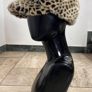 May include: A newsboy cap featuring a tan and black leopard print pattern made of a soft, fuzzy faux fur material. The cap has a rounded crown and a short, stiff brim extending from the front, displaying a textured, animal-inspired design.