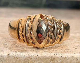 May include: A gold-colored ring with a central, faceted oval design. The ring features braided and ribbed detailing on the band, creating a textured appearance. The ring's design is symmetrical and has a polished finish.