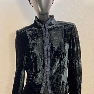 Op de afbeelding: Zwarte fluwelen jas met een hoge kraag en ruches aan de voorkant. De jas heeft lange mouwen en een getailleerd silhouet. Een decoratieve hoofdband met zilveren accenten staat op de mannequin.