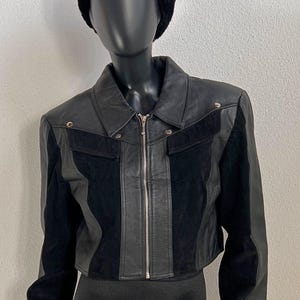 Puede incluir: Chaqueta de cuero negra con diseño corto. La chaqueta presenta un cuello, una cremallera frontal y paneles de ante y cuero. Un sombrero de piel negro está en la cabeza del maniquí. La chaqueta se exhibe en un maniquí negro.