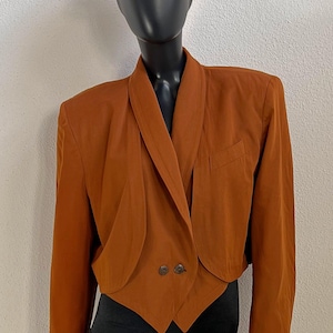 Puede incluir: Una blazer corta de color naranja quemado con cuello chal y cierre de dos botones. La blazer se exhibe en un maniquí, combinada con un sombrero de paja. La chaqueta tiene un bolsillo en el lado izquierdo.