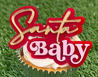 Könnte beinhalten: Ein roter und goldener Cupcake-Topper mit den Worten "Santa Baby" in einer kursiv geschriebenen Schrift. Der Topper befindet sich auf einem weißen Cupcake mit einer grünen Cupcake-Verkleidung.
