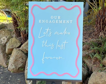 May include: A light blue sign with a pink wavy border reads "Our Engagement Let's make this last forever..." The sign is on a black easel.