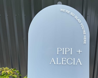 May include: A light blue arch-shaped sign with white text that reads "We're so glad you're here", "Pipi + Alecia", and "24 April 2024". The sign is on a black easel.