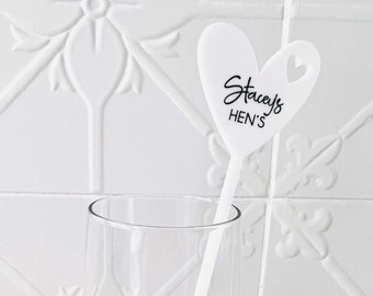 May include: White heart-shaped plastic champagne flute charm with the text "Stacey's HEN'S" in black lettering. The charm is attached to a clear plastic stick.