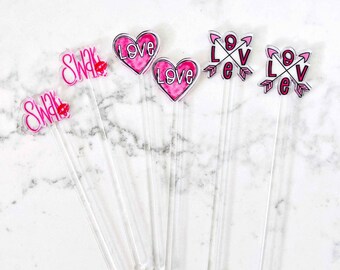 May include: Six clear acrylic drink stirrers with pink and white designs. Two stirrers have a pink heart with the word "Love" written on them. Two stirrers have a pink and white design with the word "Love" written with arrows. Two stirrers have a pink lip with the word "Swak" written on them.