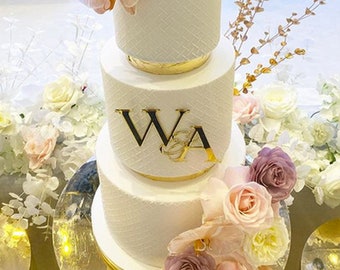 Puede incluir: Un pastel de bodas blanco con detalles dorados y las iniciales "W & A" en el nivel intermedio. El pastel está decorado con flores rosas y moradas.