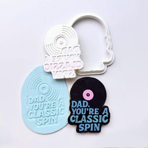 May include: A collection of cookie cutters and a cookie in the shape of a vinyl record. The cookie cutters are white and the cookie is black with a pink center. The text "DAD, YOU'RE A CLASSIC SPIN" is on the cookie.