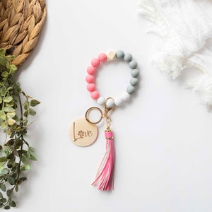 May include: A pink, gray, and white beaded keychain with a wooden charm that says "Love" with a paw print. The keychain has a gold key ring and a pink tassel.
