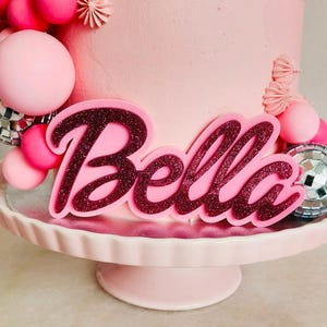 Könnte beinhalten: Rosafarbener Kuchen mit rosa und silbernen Dekorationen. Der Kuchen ist mit dem Namen "Bella" in rosa glitzernden Buchstaben verziert.