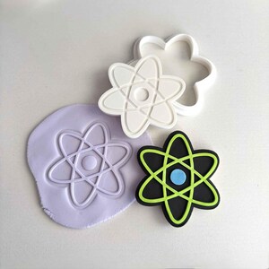 May include: A collection of atom-shaped cookie cutters and a matching impression on purple dough. The cutters are white and the atom design is also featured in black with green and blue accents. The cutters are for baking.