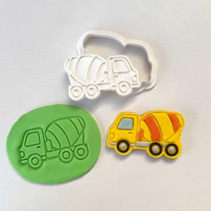 Może przedstawiać: Biała plastikowa wykrawaczka do ciastek w kształcie betoniarki obok zielonego odcisku z plasteliny oraz żółto-pomarańczowej betoniarki. Żółta ciężarówka ma czarne koła.