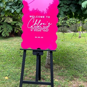 Könnte beinhalten: Rosa Willkommens-Schild mit weißem Text "Willkommen bei Chloe's Hens 04.02.23" auf einem schwarzen Staffelei.