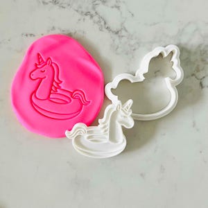 Può includere: Un impasto rosa con un'impronta di unicorno, accanto a due formine bianche a forma di unicorno. Il design dell'unicorno presenta un salvagente. Le formine sono a forma di unicorno.