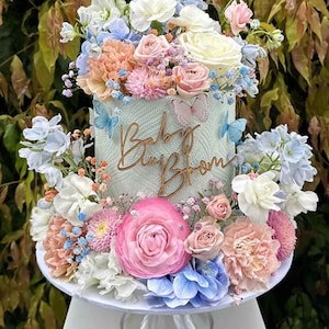 Może przedstawiać: Jasnoniebieski tort ozdobiony różnymi kwiatami, w tym różowymi, białymi i niebieskimi. Tort jest ozdobiony drewnianym napisem "Baby Bloom".