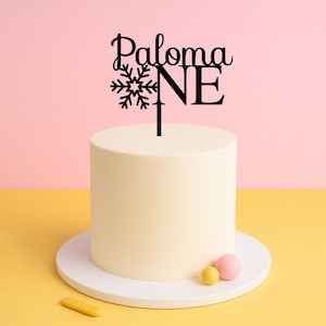 May include: Black cake topper with a snowflake and the words "Paloma ONE" for a first birthday party.