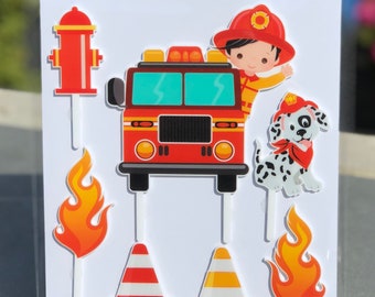 May include: A set of red, yellow, and white acrylic cake toppers featuring a fire truck, a fireman, a dalmatian dog, flames, and traffic cones. The cake toppers are on a white background with the text "Designer Cake Topper Decorating Kit" and "FRAGILE take care when removing item from backing board".
