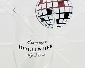 May include: A clear champagne flute with the text "Champagne BOLLINGER Ay-France" printed in black. A round, decorative piece with a globe design in white and brown is attached to the rim of the glass.