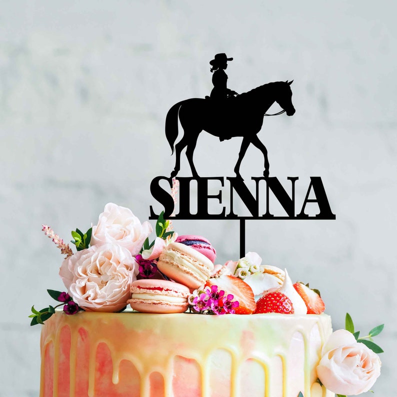 May include: A black silhouette of a girl riding a horse with the name "SIENNA" on a cake decorated with pink and white flowers, macarons, and strawberries.