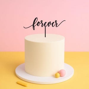 May include: A white cake with a black "forever" topper on top. The cake is on a white cake stand and there are two small round decorations on the stand, one yellow and one pink.