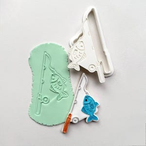 Op de afbeelding: Een set koekjessnijders met visthema. Eén is van wit plastic, een andere een groene klei-afdruk en de derde een blauwe vis met een oranje hengel. De uitstekers zijn ontworpen om visvormige koekjes te maken.
