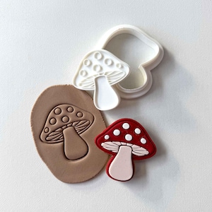 Puede incluir: Un cortador de galletas en forma de hongo blanco junto a una galleta de hongo roja y blanca y una impresión de arcilla marrón de un hongo. La galleta de hongo tiene una tapa roja con puntos blancos y un tallo blanco.