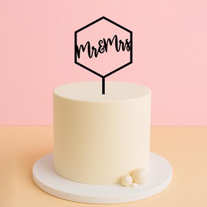以下が含まれることがあります： 白いケーキに、黒い六角形のケーキトッパーが乗っています。トッパーには、筆記体で「Mr & Mrs」と書かれています。