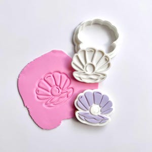 May include: A white plastic seashell-shaped cookie cutter and a matching stamp, alongside a pink clay impression and a purple seashell-shaped cookie. The design features a pearl in the center of the shell, ideal for crafting or baking.