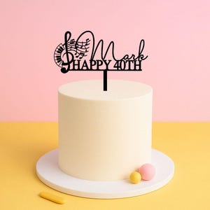 Op de afbeelding: Een witte cake met een zwarte caketopper waarop "Mark Happy 40th" staat met een solsleutel en noten. De cake staat op een witte cake standaard met twee kleine ronde snoepjes op de standaard.