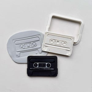 May include: A collection of cassette tape-shaped cookie cutters in various stages. One is a gray imprint, another is a white cutter, and the last is a black cutter. The cutters are designed to create retro-themed cookies.