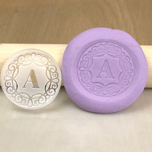 Könnte beinhalten: Ein klarer Acrylstempel mit einem dekorativen Rand und dem Buchstaben "A" in der Mitte. Der Stempel wird neben einer lilafarbenen Silikonform mit dem gleichen Design gezeigt.