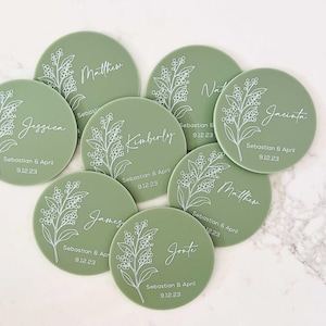 May include: Ten sage green round place cards with white floral designs and names printed in white. Each card has the date "Sebastian & April 912.23" printed on it.