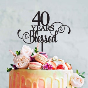 Peut inclure: Un topper de gâteau noir avec le texte "40 Years Blessed" dans une police d'écriture cursive décorative. Le topper est placé sur un gâteau blanc et rose décoré de macarons, de fraises et de fleurs.
