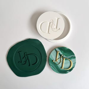 Könnte beinhalten: Ein weißer runder Stempel mit den Initialen "J&D" und ein dunkelgrünes Wachssiegel mit denselben Initialen. Ein runder, marmorierter grün-goldener Entwurf mit den Initialen "J&D" wird ebenfalls gezeigt.