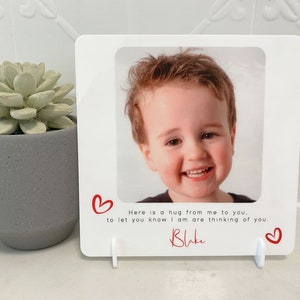 May include: White photo frame with a picture of a young boy smiling. The frame has a red heart on each side and the text "Here is a hug from me to you, to let you know I am are thinking of you. Blake" at the bottom.