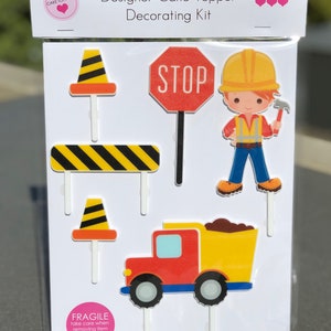 May include: Construction themed cake topper set with a yellow dumper truck, a stop sign, a construction worker, and two traffic cones. The set is on a white background with the text "Designer Cake Topper Decorating Kit".