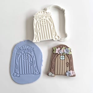 May include: A fairy door cookie cutter and two examples of the finished product. The cutter is white plastic. One finished door is brown with a floral crown and the other is a blue impression.