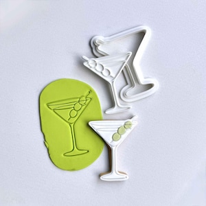 May include: White plastic cookie cutters shaped like martini glasses with olives. One martini is cut out of green dough. A finished cookie is shown, along with a cookie cutter.
