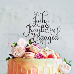 May include: A white cake with pink and yellow frosting and a black acrylic cake topper that says "Josh & Kaylie Engaged" in a cursive font. The cake is decorated with pink roses, macarons, and strawberries.
