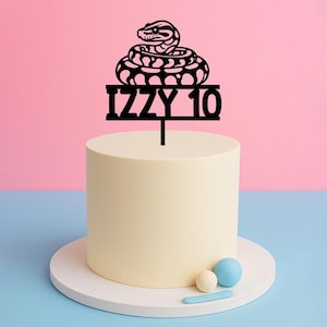 May include: A cake with a black snake cake topper that reads "IZZY 10". The cake is light yellow with a white base. The background is pink and blue. There are three small decorative balls on the base of the cake.
