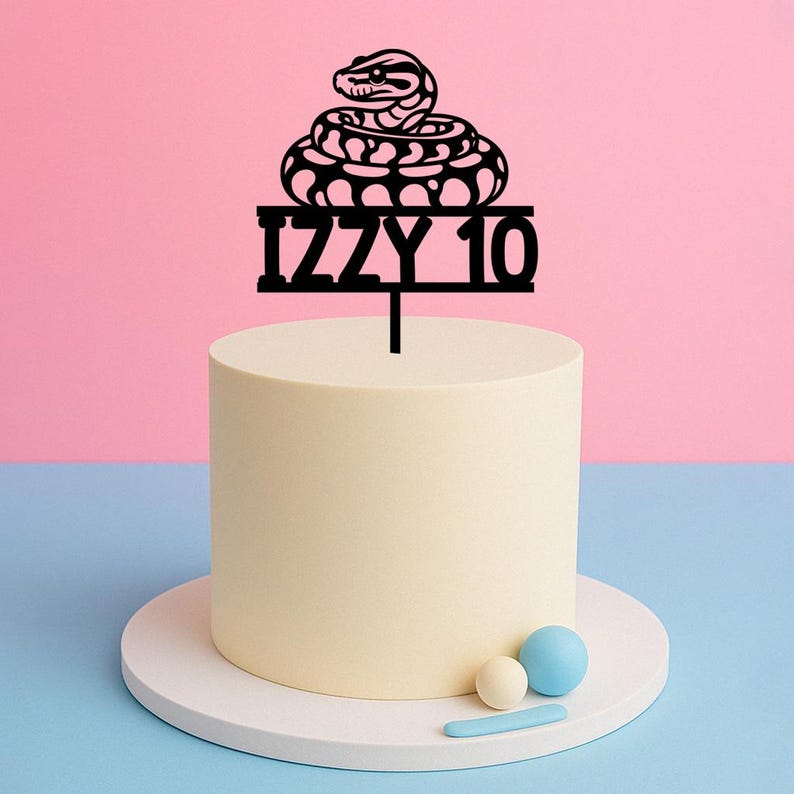 Peut inclure: Un g&acirc;teau jaune clair avec un cake topper noir en forme de serpent avec l'inscription "IZZY 10". Le g&acirc;teau est pos&eacute; sur un socle blanc, sur fond rose et bleu. Trois petites boules d&eacute;coratives sont sur la base du g&acirc;teau.
